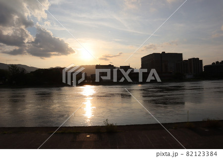 雲で中でオレンジ色に光る太陽と灰色の雲と色づく薄雲と太陽の道の川面の川と山と逆光で暗く見える町の風景 雲で中でオレンジ色に光る太陽と灰色の雲と色づく薄雲と太陽の道の川面の川と山と逆光で暗く見える町の風景 82123384