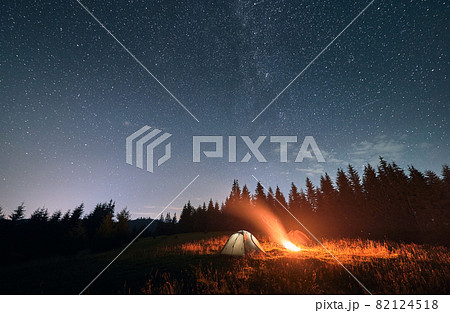 Night camping under amazing starry sky. Wide angle view on beautiful landscape in mountains. Two tourist tents and campfire, wall of spruces on background. Concept of travelling and astrophotography Night camping under amazing starry sky. Wide angle view on beautiful landscape in mountains. Two tourist tents and campfire, wall of spruces on background. Concept of travelling and astrophotography 82124518