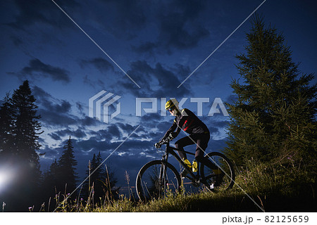 Man cyclist riding bicycle downhill with beautiful blue evening sky on background. Male bicyclist in sports cycling suit cycling down grassy hill at night. Concept of sport, biking and active leisure. 82125659