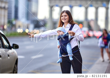 Woman with child in baby sling stops taxi on busy city street. 82131191