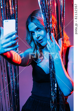 Christmas selfie. Beautiful woman. Festive look. Happy stylish lady black dress curly hair making photo mobile phone shimmering cascade curtain red blue neon light. 82132567