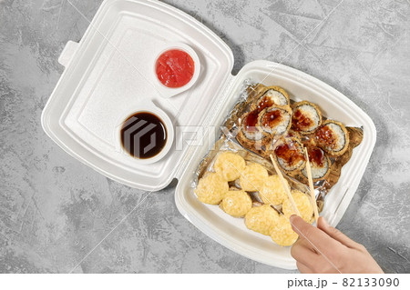 Delicious sushi rolls in disposable plastic boxes. The hand takes a roll with chopsticks. Food delivery service concept, flat stacking 82133090