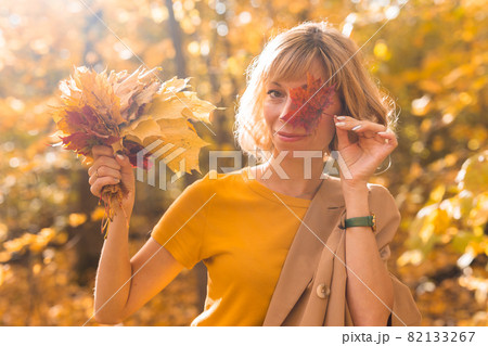 Young blonde woman covering one eye with red maple leaf. Autumn and season concept. Outdoor fall female portrait close-up with foliage 82133267
