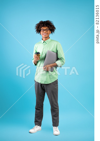 Modern student. Happy black guy with laptop computer and takeaway coffee posing to camera on blue background Modern student. Happy black guy with laptop computer and takeaway coffee posing to camera on blue background 82136015