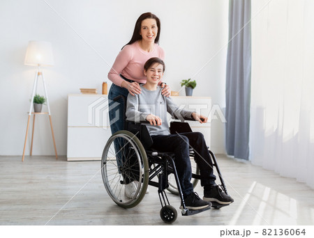 Full length portrait of happy young mother and her disabled teen son in wheelchair looking at camera, posing indoors 82136064