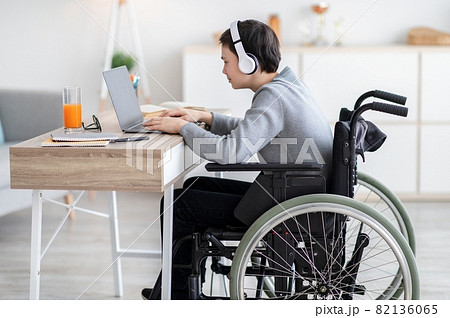 Side view of handicapped teen boy in wheelchair wearing headphones, having video chat with teacher or classmate at home 82136065