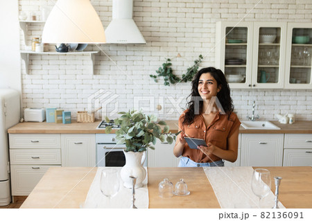 Cheery young brunette woman using tablet computer, browsing internet or social media, working online in kitchen Cheery young brunette woman using tablet computer, browsing internet or social media, working online in kitchen 82136091