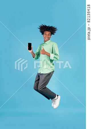Cool mobile app. African american guy jumping and showing smartphone with blank screen, gesturing thumb up 82136418