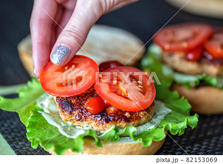 A woman's hand lays down a slice of tomato on a delicious homemade burger. Handmade homemade burgers, close-up A woman's hand lays down a slice of tomato on a delicious homemade burger. Handmade homemade burgers, close-up 82153069