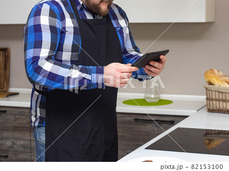 A man in the kitchen is watching a recipe for cooking a dish on a tablet on the Internet, background 82153100