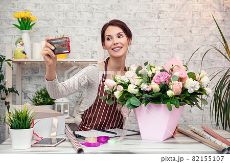 Young woman florist at work. Business woman takes a selfie with a bouquet, happy smiling woman making bunch at flower shop. Beautiful girl florist with flowers. Female florist working in from home 82155107