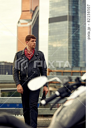 Portrait of stylish male dressed in a black leather jacket and bandana sitting on his custom-made retro motorcycle, looking away, buildings of the big city on background. Portrait of stylish male dressed in a black leather jacket and bandana sitting on his custom-made retro motorcycle, looking away, buildings of the big city on background. 82156507