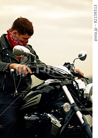 Portrait of stylish male dressed in a black leather jacket and bandana sitting on his custom-made retro motorcycle, looking away, buildings of the big city on background. 82156513