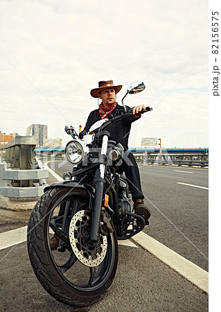 Portrait of stylish male dressed in a black leather jacket and bandana sitting on his custom-made retro motorcycle, looking away, buildings of the big city on background. 82156575