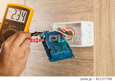 Electrician using a digital meter to measure the voltage at a wall socket on a wooden wall. 82157766