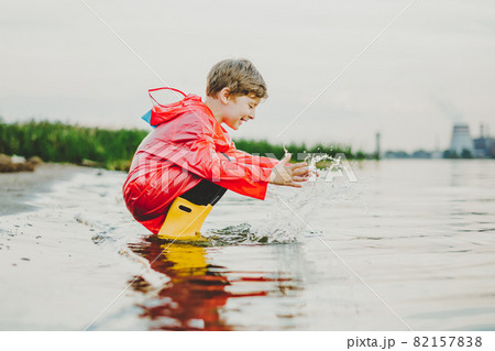 Boy in a red raincoat and yellow rubber boots playing with water at the beach. School kid in a waterproof coat touching water at sea. Child having fun with waves at the shore 82157838