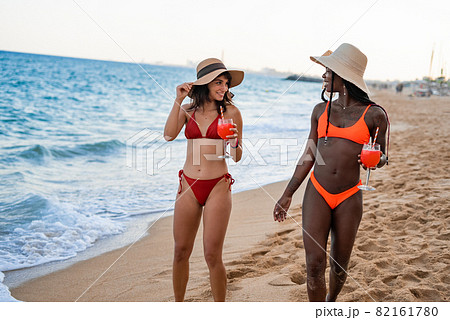 Young fit multiracial girlfriends in colorful swimwear and straw hats with glasses of refreshing beverages smiling and looking at each other while strolling together on sandy beach during summer 82161780