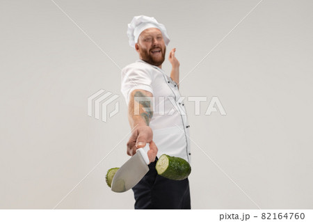 Unique cutting of vegetables. One hadsome bearded man, cook, male chef in white uniform cut cucumber isolated on white background. Unique cutting of vegetables. One hadsome bearded man, cook, male chef in white uniform cut cucumber isolated on white background. 82164760