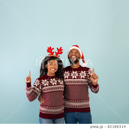 Holiday offer. Happy black spouses in identical Christmas sweaters pointing up at copy space, blue background 82166314
