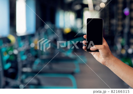 Male Hand Holding Smartphone With Blank Black Screen Over Gym Interior Background Male Hand Holding Smartphone With Blank Black Screen Over Gym Interior Background 82166315