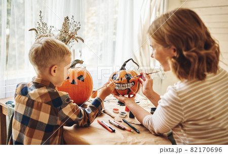 Smiling mother and child boy drawing scary faces on Halloween pumpkins while sitting in cozy kitchen Smiling mother and child boy drawing scary faces on Halloween pumpkins while sitting in cozy kitchen 82170696