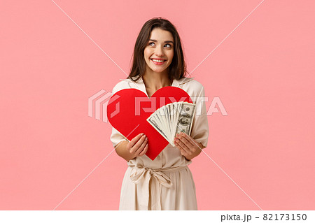 Love, tenderness and relationship concept. Cute and silly girl dreaming of wealthy rich man and true love, holding money dollars and heart card, smiling looking up imaging thing, pink background 82173150