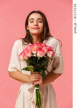 Full-length vertical portrait romantic and dreamy, sensual brunette girl in dress, holding beautiful bouquet flowers, smelling roses with closed eyes and smiling, standing happy over pink background 82173156