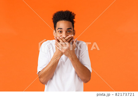Guy made funny prank trying hold laugh. Hilarious and amused african-american male in white t-shirt, giggle cover mouth with palms and staring amazed camera, chuckling, standing orange background Guy made funny prank trying hold laugh. Hilarious and amused african-american male in white t-shirt, giggle cover mouth with palms and staring amazed camera, chuckling, standing orange background 82173721