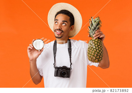 Happy dreamy handsome male tourist enjoying vacation, looking at bright sun partying on beach, holding coconut and pineapple, making cocktail, standing orange background with camera 82173892