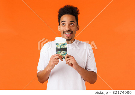 Dreamy cheerful, smiling african-american guy with moustache, afro haircut, holding passport with flight tickets, looking up grinning joyfully, picturing himself on luxury resort, orange background 82174000