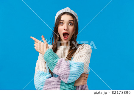 Shocking low prices, check this out. Astonished and speechless, startled excited young woman with long dark hair, winter sweater and hat, open mouth fascinated, pointing finger left, blue background 82178896