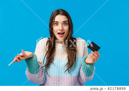 Surprised and wondered, amused cute caucasian woman in winter sweater, shrugging amazed, holding credit card and smartphone, got cashback, standing blue background joyful 82178899