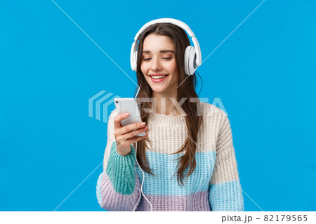 Technology, millennials and lifestyle concept. Carefree cute brunette female student put on headphones, plug-in smartphone picking song and smiling, standing blue background make playlist for study 82179565