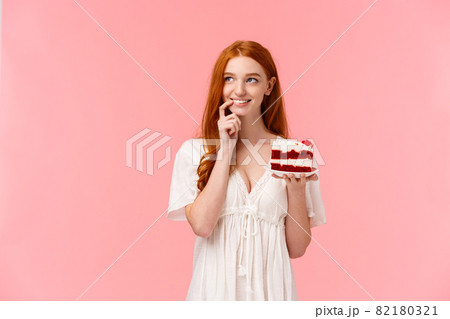 Lovely, sensual caucasian redhead girl with silly smile, thinking, dreaming about something, touch lip and gazing up thoughtful as holding plate with delicious cake, pink background 82180321