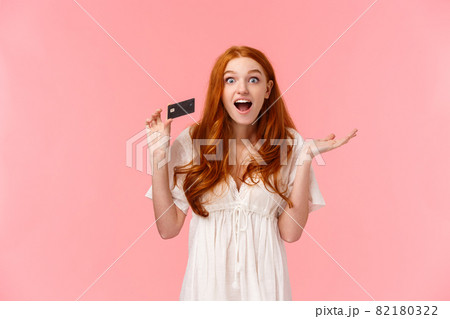 Waist-up portrait amused, wondered and excited fashionable feminine redhead girl telling about great promos and bonuses in bank, holding credit card and smiling astonished, pink background 82180322