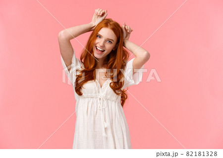 Charming, pretty and cute redhead female mimicking dog, holding hands over head as puppy ears, tilt head and smiling, playfully fool around, feeling goofy, standing pink background kawaii pose 82181328