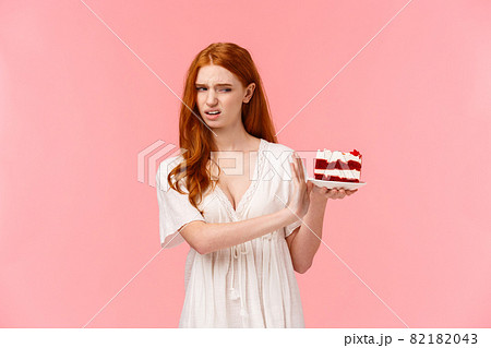 Take it away, gross. Ignorant and picky spoiled redhead birthday girl refusing eating dessert, holding plate with cake and making stop, rejection gesture, turn away with disgust, pink background 82182043