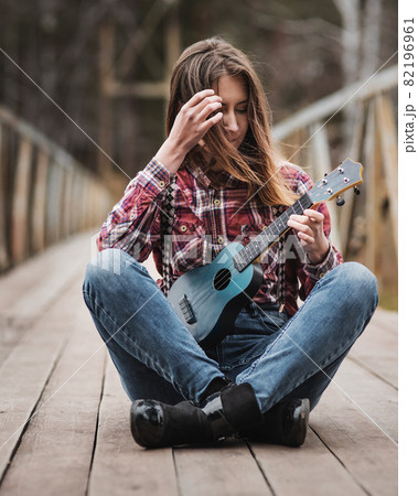 Young woman sitting and playing ukulele guitar on bridge in autumn forest Young woman sitting and playing ukulele guitar on bridge in autumn forest 82196961