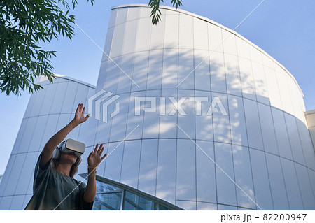 A girl is having fun in virtual reality glasses, standing against the background of a mirrored building. The concept of the future. High-quality photo 82200947