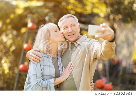 Senior couple spending time in sunny garden together, taking selfies, woman kissing husband in cheek Senior couple spending time in sunny garden together, taking selfies, woman kissing husband in cheek 82202933