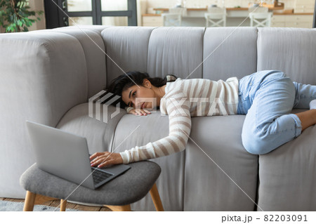 Tired young Caucasian woman lying on couch, using laptop, too exhausted or bored for online work or studies at home 82203091