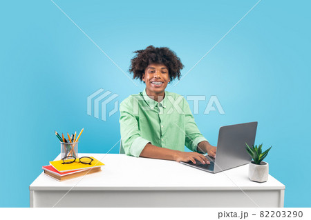 Happy african american teen guy sitting at desk and using laptop, doing homework, studying online, blue background Happy african american teen guy sitting at desk and using laptop, doing homework, studying online, blue background 82203290