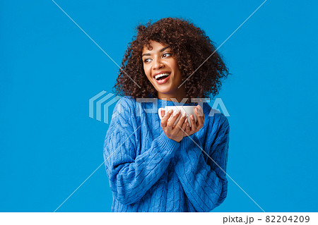 Girl having chat with girlfriend while drinking coffee. Cheerful and cute african-american lovely woman with afro haircut, in sweater, tilt head gazing left and holding hot cup delicious tea 82204209