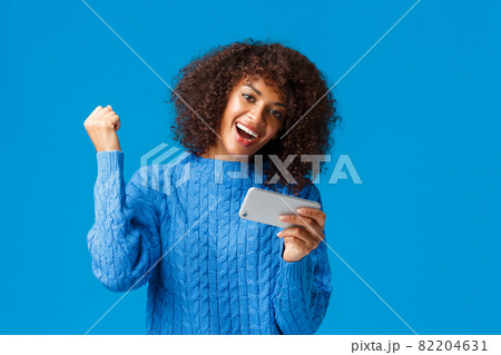 Excited happy lucky african-american woman passed level, finished race first in game, holding smartphone playing app, cheering, fist up like champion, celebrating victory, blue background 82204631