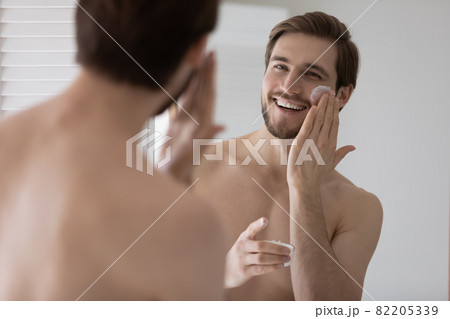 Handsome young man looks in mirror apply facial cream Handsome young man looks in mirror apply facial cream 82205339