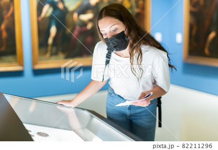 Young girl in a protective mask in the museum looks at the exposition behind a glass case 82211296