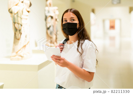Young girl wearing a protective mask in the museum looks at the exhibits 82211330