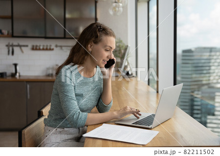 Young Caucasian woman talk on smartphone working on computer Young Caucasian woman talk on smartphone working on computer 82211502
