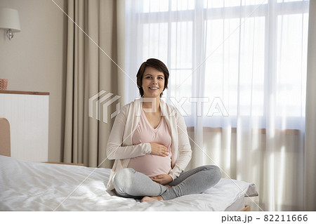 Excited young future mom sit cross legged on comfortable bed 82211666