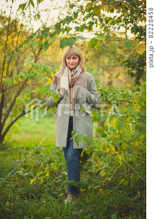 A blonde woman with a short haircut walks through the forest in a gray wool coat and a plaid scarf. Walking in nature in autumn forest A blonde woman with a short haircut walks through the forest in a gray wool coat and a plaid scarf. Walking in nature in autumn forest 82228458